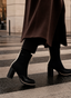 Botines de mujer AZAREY en color negro con tacón ancho, caña ajustada y detalle lateral en contraste que estiliza el diseño. La imagen muestra el calzado en movimiento sobre un paso de peatones, combinado con abrigo largo y pantalón negro, resaltando su estilo urbano, elegante y actual. Un botín versátil para looks de temporada, ideal para diario, oficina o conjuntos sofisticados con un toque moderno.